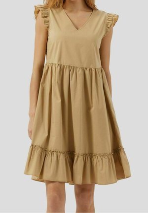 Robe beige sans manches avec décolleté en V, manches courtes volantées, taille froncée et ourlet à volants, portée par une personne debout sur un fond uni.