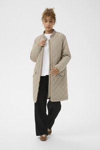 Quilttet beige lang frakke med opretstående krave, fuld lynlås og sidelommer; båret over en hvid top og sorte bukser, der viser en afslappet pasform.
