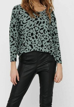 Femme portant un pull vert ample à manches longues avec imprimé léopard noir et un pantalon en cuir noir ajusté, debout devant un fond uni.