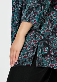 Blusa patterned con base oscura y manchas de leopardo en tonos verde azulado y marrón. Confeccionada en una tela texturada, presenta aberturas laterales y mangas sueltas.