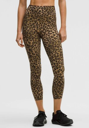 Frau trägt hoch taillierte, gekürzte Leggings mit Leopardenmuster und schwarzen Sportschuhen, steht vor einem einfarbigen Hintergrund.