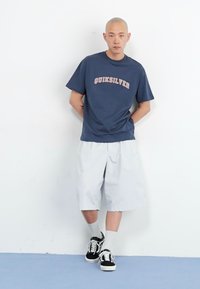 Μπλε ναυτικής απόχρωσης κοντομάνικο T-shirt με λογότυπο "Quiksilver" σε ανοιχτό ροζ, συνδυασμένο με άσπρα σορτσάκια μέχρι το γόνατο και μαύρα αθλητικά παπούτσια. Λείο υλικό.