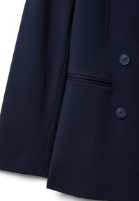 Navyblauwe stof in close-up met een deel van een colbert met twee donkere knopen en een paspelzak aan de onderrechterzijde.