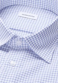 Chemise blanche à motif à carreaux bleu clair, avec un col large, deux boutons blancs et une étiquette de marque visible à l'intérieur du col.