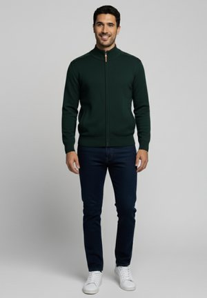 Chaqueta de punto verde con cremallera y collar alto, ajuste slim, y puños acanalados, combinada con jeans oscuros y zapatillas blancas.
