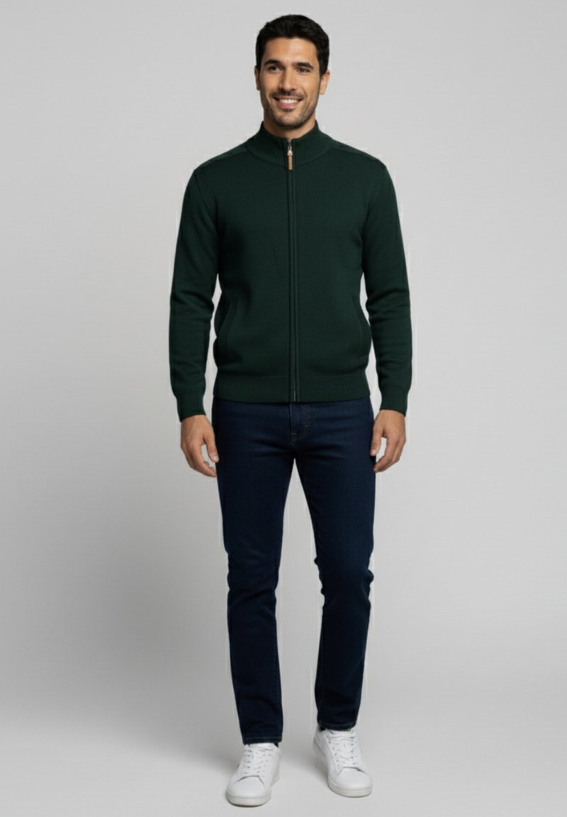 Chaqueta de punto verde con cremallera y collar alto, ajuste slim, y puños acanalados, combinada con jeans oscuros y zapatillas blancas.