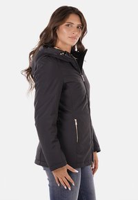 Giacca nera con zip e cappuccio, dotata di tasche frontali con zip e dettagli in metallo dorato. Texture morbida e design aderente.