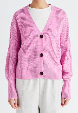 Personne portant un cardigan en maille épaisse rose avec trois boutons foncés sur un haut blanc et un pantalon gris clair.