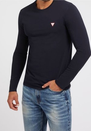 Mann trägt enganliegendes schwarzes Langarmshirt mit Guess-Logo auf der Brust und hellblaue ausgewaschene Denim-Jeans, Hand in der Tasche.