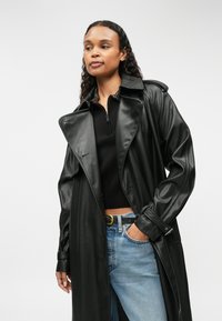 Czarny perforowany skórzany płaszcz o szerokim kołnierzu, z podwiniętymi rękawami i zaznaczoną talią. Zestawiony z czarną bluzką i dżinsami.