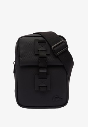 Bolso bandolera de tela negra con superficie texturizada, correa ajustable, cierre de hebilla prominente y detalle de logo grabado en la parte delantera.