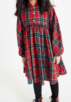 Enfant portant une robe à manches longues en tartan rouge, vert et bleu marine avec une taille froncée, des collants noirs et des chaussures à lacets rouge foncé, se tenant devant un fond blanc.