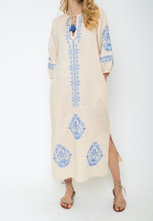 Robe midi beige avec broderie florale bleue, manches trois-quarts, fente latérale, lien noué à l'avant avec des pompons, portée avec des sandales beiges à lacets.