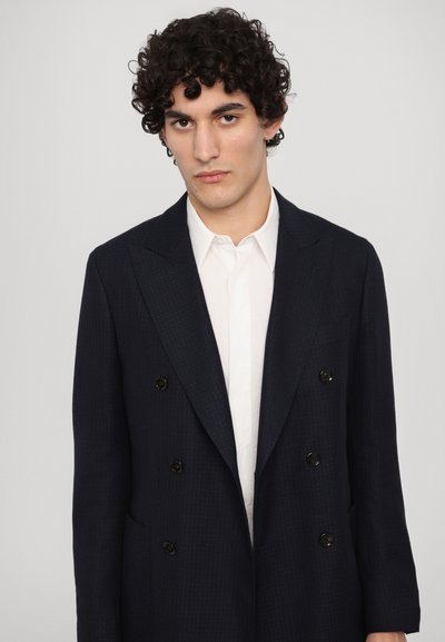 Blazer bleu marine cu două rânduri de nasturi, dintr-un material texturat, având nasturi mari negri, rever larg și o croială ajustată. Cămașă albă dedesubt.