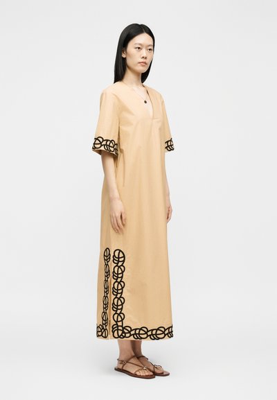 Femme portant une robe beige longueur cheville avec une broderie circulaire noire sur les manches et l’ourlet, debout en sandales couleur camel sur un fond uni.