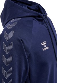 Marineblauwe hoodie gemaakt van zacht materiaal, met witte chevronpatronen op de mouwen en een logo op de borst. Klassiek ontwerp met een trekkoord.