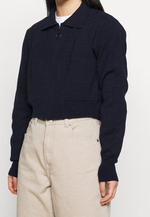 Frau trägt einen kurz geschnittenen marineblauen Reißverschluss-Pullover mit Kragen über einem weißen Hemd und hoch taillierten beigen Hosen.