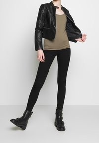 Giacca nera in pelle trapuntata, canotta oliva, jeans skinny neri e stivali neri alla caviglia con fibbie, su uno sfondo semplice.