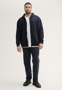Marine blauwe ritscapuchontrui, met een wit t-shirt eronder, gecombineerd met marineblauwe broek en zwarte sneakers. Zacht materiaal, ontspannen pasvorm en voorzakken.