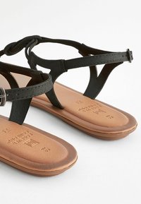 Zwarte leren sandalen met een platte, bruine rubberen zool. Kenmerken zijn een teenpost en een verstelbare enkelband met een gespsluiting.