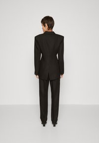 Musta muotonsa suit, jossa on leveät hartiat, viistetty kaulus ja slim fit. Takissa on keskiventti; housut ovat suorat.