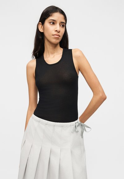 Νέα γυναίκα με μακριά σκούρα μαλλιά, που φοράει εφαρμοστό μαύρο αμάνικο τοπ και ανοιχτό γκρι πλισέ φούστα δεμένη στη μέση.