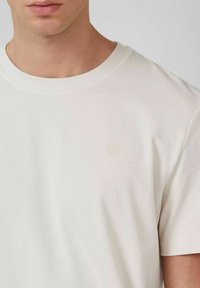 Male wearing a plain off-white crew neck short-sleeve T-shirt with a subtle small logo on the upper left chest.