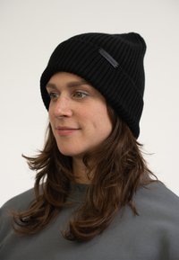 Beanie nero a coste con risvolto e design strutturato. Presenta un piccolo etichetta nera decorative sulla parte anteriore.