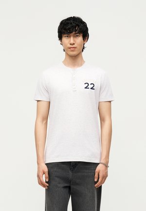 Jeune homme aux cheveux noirs bouclés portant un t-shirt henley gris clair à manches courtes avec le logo "22" et un jean foncé, debout devant un fond uni.
