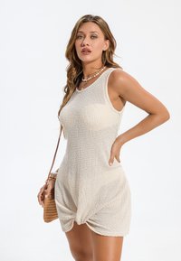 Pletené béžové šaty s tílkem, s uzlovým detailem vpředu, bez rukávů a s jemným strukturovaným vzorem. Nošeno s malou kabelkou.