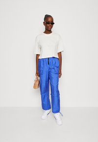 T-shirt bianco croppato, pantaloni cargo blu con vita elasticizzata e tasche, sneakers bianche con platform, holding una borsa beige. Outfit casual moderno.