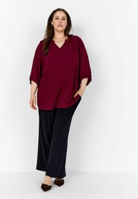 Blusa bordeaux fluida con scollatura a V e yoke arricciato, abbinata a pantaloni neri a gamba larga. L'outfit è completato da scarpe marroni.