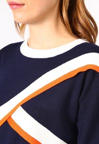 Marineblå tröja med vita och orangea diagonala ränder på framsidan. Tillverkad av ett mjukt stickat material, med en rundad halsdesign.