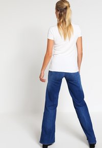 Kvinna med långt blont hår klädd i en vit kortärmad skjorta och blå vida jeans, stående med ryggen mot kameran.