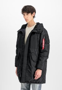 Melnā polsterētā parka ar rāvējslēdzēju un kapuci, ar priekšējiem kabatām un sānu plecu kabatu. Ievērojama sarkanā birka uz rāvējslēdzēja.