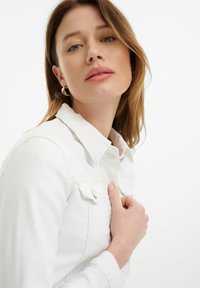 Bijela traper jakna s ovratnikom, prednjim zatvaranjem na gumbe i džepom na prsima; glatka tekstura, prilagođen dizajn; model drži jaknu.