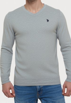 Mand iført lysegrå V-hals sweater med tekstur og lille mørkt logo på brystet samt blå jeans, stående med armene ned langs siderne.