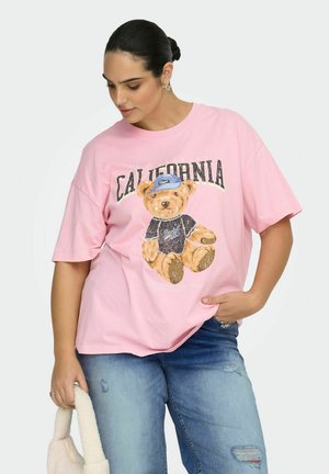 Femme aux cheveux foncés attachés en chignon, portant un t-shirt rose oversize avec un motif d'ours en peluche et le texte "California", un jean bleu déchiré, tenant un sac blanc.