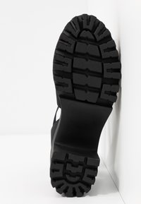 Svart gummisula med en strukturerad yta och djupa spår för bättre grepp, samt en något upphöjd häl för stöd.