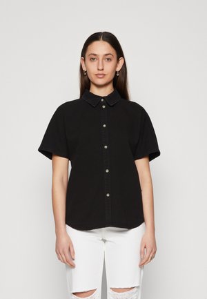 Svart kortermet skjorte med krage, knappestolpe og myk, strukturert stoff, sammen med hvite slitte denimbukser.