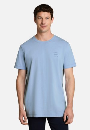 Mann mit kurzen lockigen Haaren, der ein schlichtes hellblaues kurzärmeliges T-Shirt und dunkle Hosen trägt, steht vor einem weißen Hintergrund.