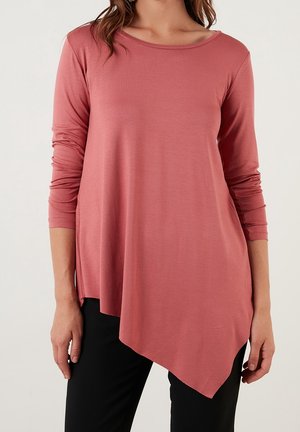 Frau trägt ein langärmliges Top in staubigem Rosé mit asymmetrischem Saum und schwarze Hose, steht vor einem schlichten hellen Hintergrund.