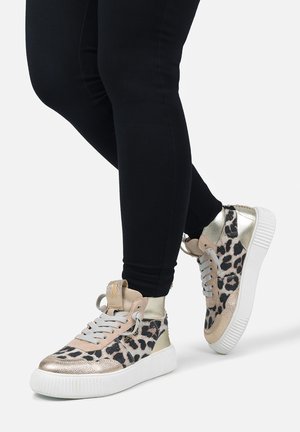 Person iført høje sneakers i metallisk guld og leopardprint med hvide såler og lysegrå snørebånd, kombineret med sorte leggings.
