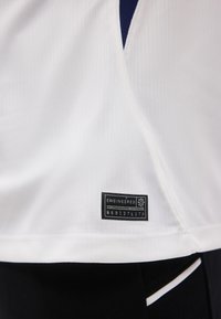 Tissu côtelé blanc avec une étiquette rectangulaire noire portant la mention « ENGINEERED » et un code, cousue près de l'ourlet sur un vêtement noir et blanc en dessous.