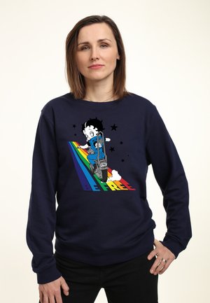 Kvinde iført en marineblå sweater med en tegnefilmsfigur, der kører på motorcykel over regnbuestriber, og teksten "VÆR FRIT" nedenfor.