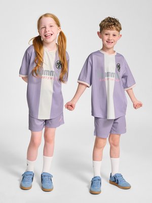 Twee glimlachende kinderen staan naast elkaar hand in hand, gekleed in bijpassende paarse en witte sportkleding met blauwe sneakers.