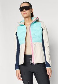 Lichte jas met een turquoise gewatteerde bovenkant, crèmekleurige en marineblauwe zijkanten, met een roze rits en decoratieve lintdetails.
