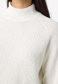 Pleteni pulover u boji slonovače s visokim rebrastim ovratnikom, koji se može pohvaliti teksturiranim uzorkom i raglan rukavima. Mekani, udobni materijali pružaju toplinu.