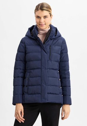 Marineblaue Steppjacke mit Kapuze, ausgestattet mit einer Reißverschluss-Brusttasche, seitlichen Knöpfen und gesteppter Struktur. Tragen über einem hellen Rollkragenpullover.