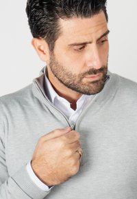 Pull zippé gris clair en matière tricotée, doté d'un col et de poignets côtelés, porté par-dessus une chemise blanche à col.